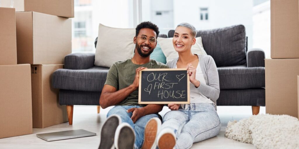 Happy couple sitting on the floor of their new home, holding a sign that reads 'Our First House,' surrounded by moving boxes.