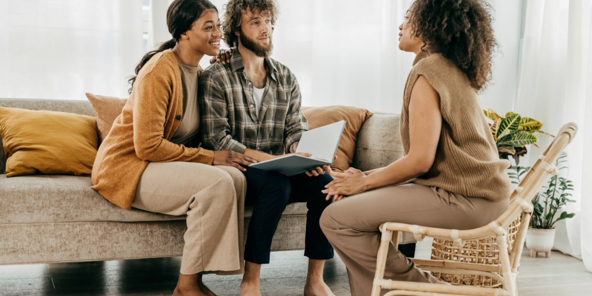 Smiling couple sitting on a couch, attentively listening to a realtor seated across from them in a cozy, modern living room.