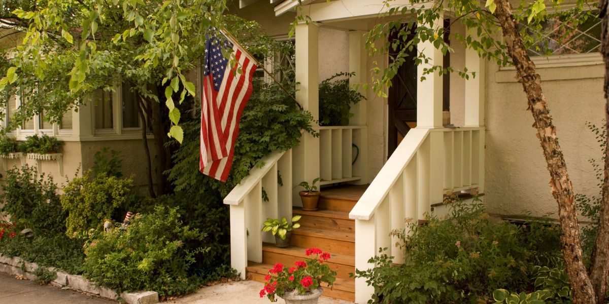 american flag home Front porch of a house with an American flag, surrounded by greenery and potted plants