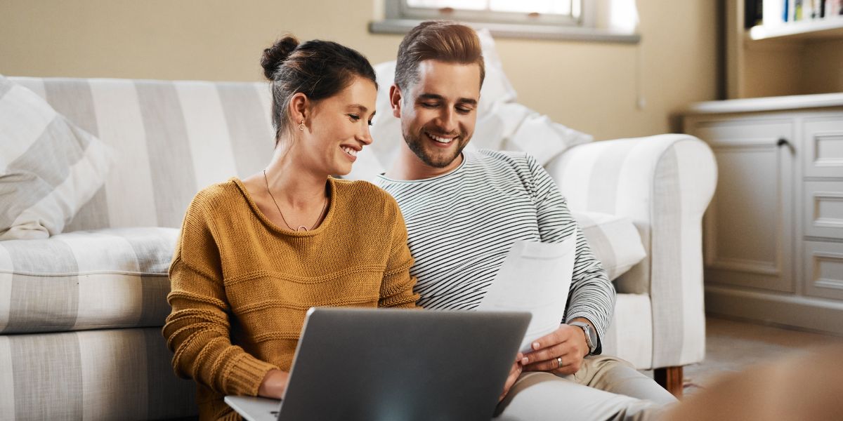 A couple sitting on the floor with a laptop and paper documents, smiling and reviewing credit scores together.