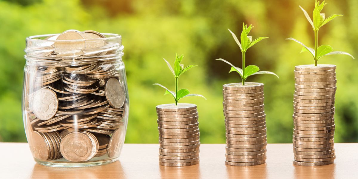 Three stacks of coins with green plants growing on top, next to a jar filled with coins, symbolizing savings growth