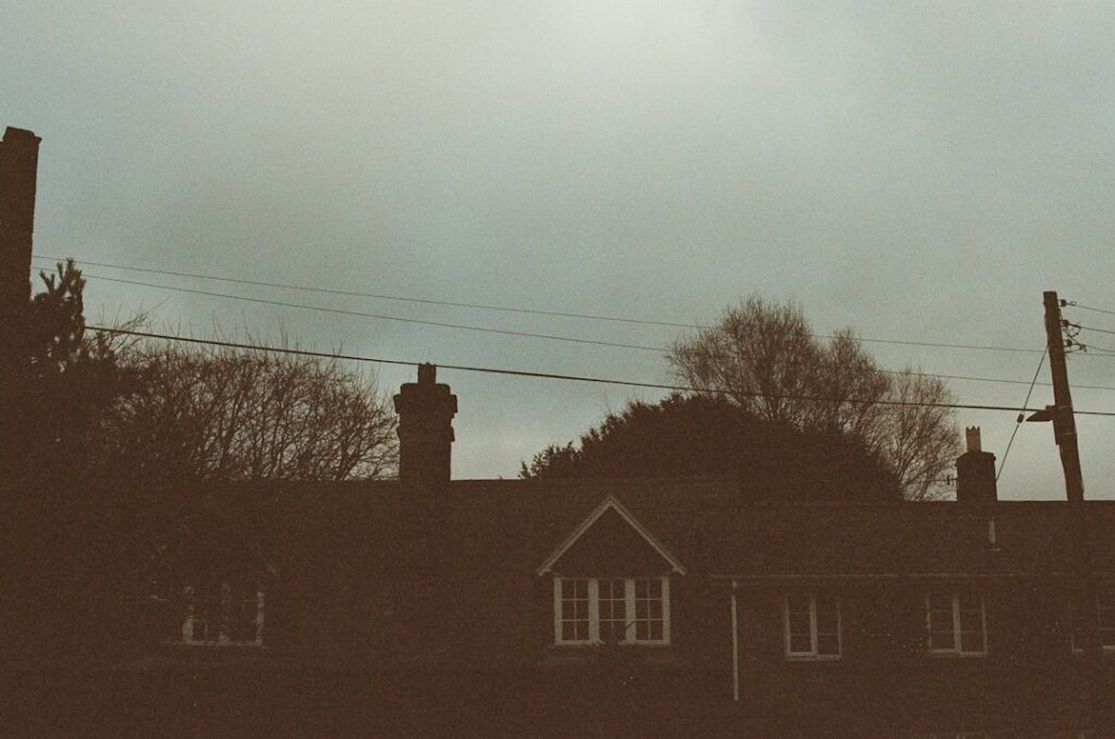 Sturdy suburban house with a well-maintained roof stands under a dark, dramatic sky filled with gathering storm clouds, sy...